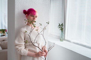 young woman with pink hair holding tree branch with first leaves in modern appartment at springtime