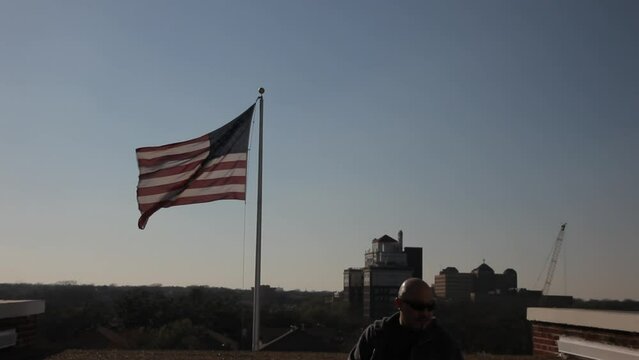 Pan down to middle aged man sitting on rooftop ledge near American flag