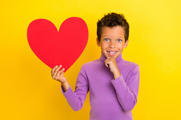 Photo of positive cute boy arm touch chin hold red paper heart card isolated on yellow color background