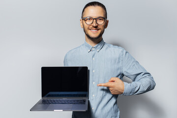 Studio portrait of young smiling man, holding laptop in hand, pointing index finger on screen, on white background. Wearing blue shirt and eyeglasses.