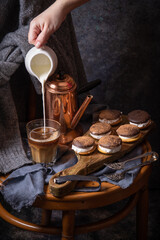 Female hand pouring milk in the coffee glass. Coffee, cakes and cozy sweater in dark style as autumn season concept. Copy space