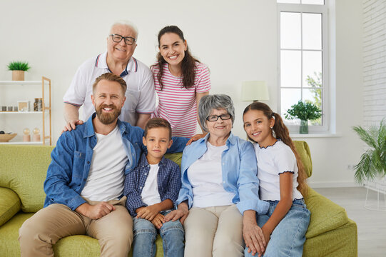 Cheerful happy family of three generations posing for family picture sitting on sofa in living room. Portrait of joyful positive grandparents with their children and grandchildren smiling at camera.