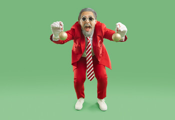 Happy man in funky costume having fun at Christmas party. Funny bearded guy in red suit, striped tie and star shaped disco glasses standing on green studio background and showing Christmas tree balls