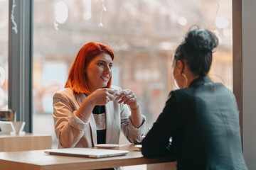 Two young business women sitting at table in cafe. Girl shows colleague information on laptop screen. Girl using smartphone, blogging. Teamwork, business meeting.