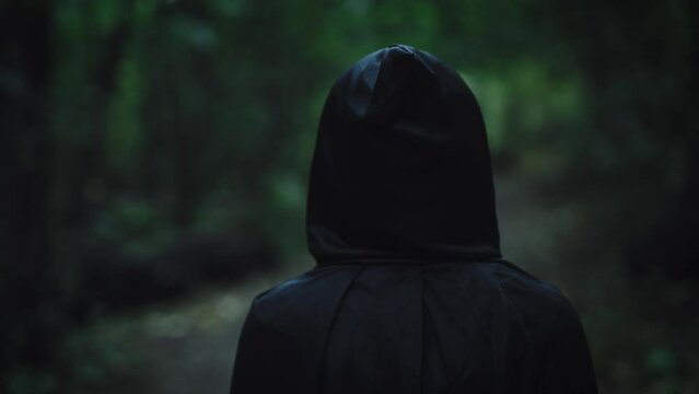 A dark figure in a black cloak with a hood walks through the enchanted forest. An unrecognizable witch from the Middle Ages walks along the path.