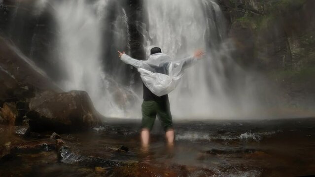 Happy man in a transparent raincoat stands with hands up at the foot of a waterfall in norway near a rock