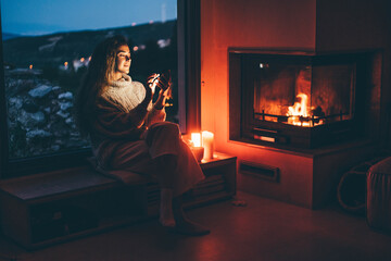 Cozy home. Young woman using phone near the fireplace.