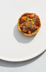 Photo of a delicious modern style handmade mini tart tartlet fulled with differ nuts like pistachio and hazelnut with salted caramel sause on a modern porcelain white flat plate on white background
