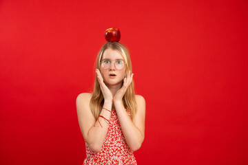 Open eyed teen girl stand with apple lying on head, empty red background, copy space. Portrait of clever child in glasses and floral print dress with surprise emotion. Idea, shock, science symbol