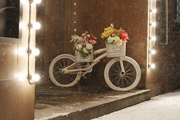 White bicycle with flowers in baskets stands at the entrance to the building in snowy weather
