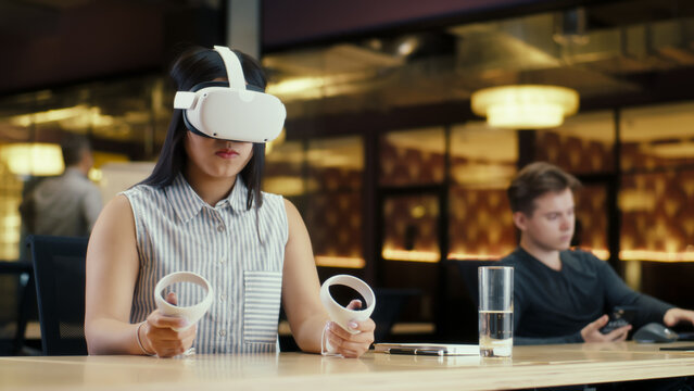 Asian female office worker using VR headset and wireless controllers, watching data in virtual reality. Work in modern office of hi-tech company with diverse colleagues. Cyberspace digital technology - Powered by Adobe