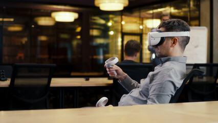 Back view of man wearing VR headset and using wireless controllers, watching data in virtual reality. Work in modern office of hi-tech company with diverse coworkers. Cyberspace digital technology