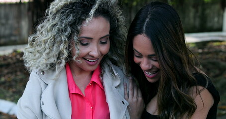 Fototapeta premium Two female friends laughing together at funny online content on cellphone device outside, real life laugh and smile
