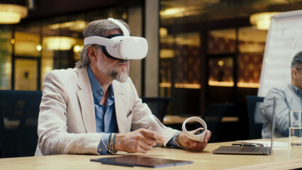 Businessman in VR headset using wireless controllers and laptop, gesturing, watching data in virtual reality. Work in modern office of hi-tech company. Cyberspace digital technology