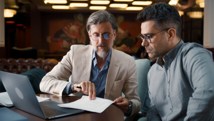Mature man using laptop, talking with serious businessman, showing papers, documents. Business...