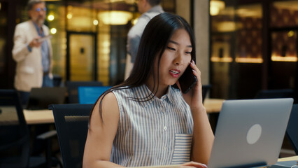 Asian woman talking with client by phone while working in modern office near colleagues, using laptop and digital tablet. Business woman discussing e-commerce startup. Strategy of the company