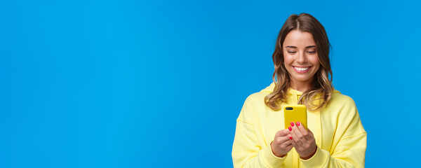 Close-up portrait silly beautiful blond european woman in yellow hoodie, smiling at mobile phone...