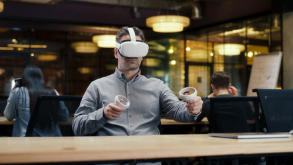 Male office worker wearing VR headset and using wireless controllers, gesturing, watching data in virtual reality. Work in office of hi-tech company near coworkers. Cyberspace digital technology