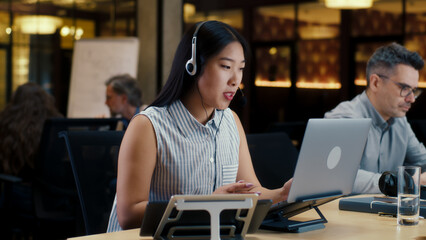 Asian woman in headset talking with client, using laptop and digital tablet. Female operator working in call center office of marketing company