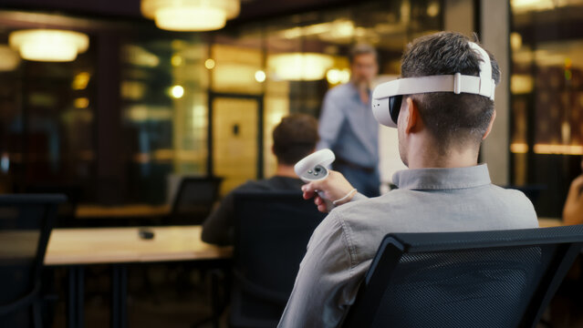 Back view of man wearing VR headset and using wireless controllers, watching data in virtual reality. Work in modern office of hi-tech company with diverse coworkers. Cyberspace digital technology