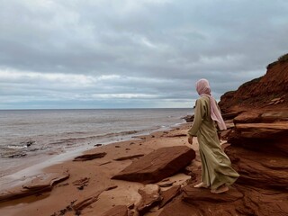 Woman in a robe with hijab standing on the rocky beach of Cavendish, Prince Edward Island, Canada
