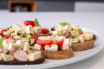 italian bread with greek cheese, tomatoes, cucumbers, radishes and basil and with various spices, on a white plate and a white kitchen table.