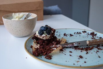 On a white table, a plate with a bite of cake and a white bowl with cream cheese frosting. Food remains in the kitchen and a blurred background.