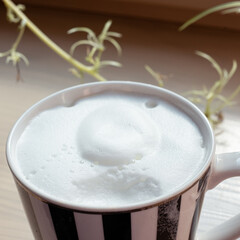 black and white striped coffee mug with coffee and milk foam on a wooden windowsill on a sunny summer morning. A sip taken.