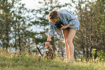 Young woman holding photo camera plays with domestic cat getting lost on empty forest glade. Female...