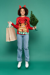 african american woman in festive outfit levitating with shopping bags and small christmas tree on green background