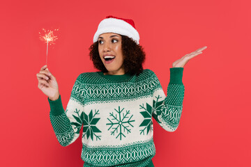 amazed african american woman in santa hat holding sparkler and pointing with hand isolated on red