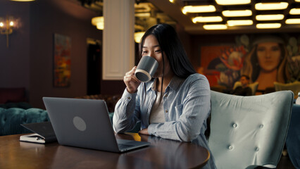 Asian businesswoman looking at screen of laptop, watching business training, working online on startup project, drinking coffee in office cafe. Worker of E-commerce marketing company