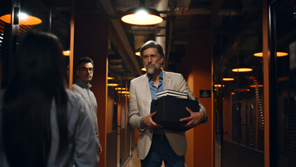 Fired male worker in suit standing in office corridor, holding box with books and personal stuff. Upset man leaving marketing company