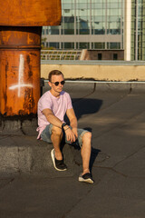 A handsome guy in denim shorts and a pink t-shirt poses beautifully in the city