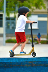 child rides a Kick scooter. playground for riding on Kick scooter. boy in a white T-shirt , hardhat and shorts riding a scooter in skatepark. young novice athlete spends free time in extreme sports.