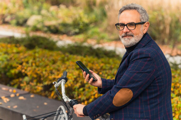 Technology in the life of elderly people. Handsome caucasian gray-haired bearded man in black-frame glasses holding his smartphone and holding bicycle wheel. High quality photo