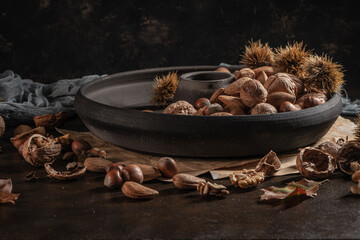 Modern design black ceramic bowl with walnuts, hazelnuts, almonds, chestnut hedgehogs on dark countertop and background. Autumn table