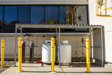 Exterior two white propane tanks and two compressed gas cylinders connected to gas distribution manifold under a metal shelter, bollards, daytime, sunny, nobody