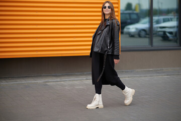 Stylish beautiful pregnant girl walks along a city street.Wearing a black leather jacket, a black long dress and white high boots and black sunglasses.