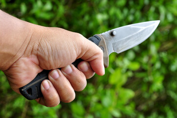 Left hand holding survival knife with green leaves blur background - obrazy, fototapety, plakaty