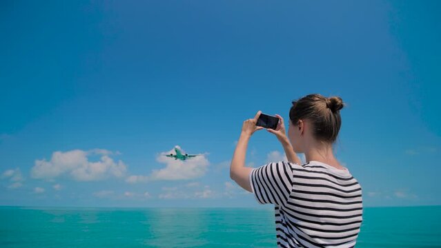 Woman holds smartphone, takes photo or shoots video of arriving passenger airplane, airliner against the blue sky - back view, wide angle view. Photography, plane spotting and technology concept