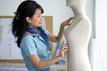 Fashion designer room. Young asian dressmaker in striped scarf use tape measure, measuring chest size of the mannequin.