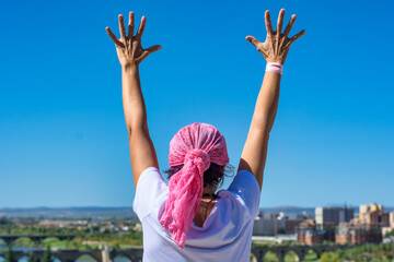 Breast Cancer Awareness Day Concept. Rear view of a woman with raised hands and pink headscarf looking up at the blue sky. Concept of overcoming. - obrazy, fototapety, plakaty