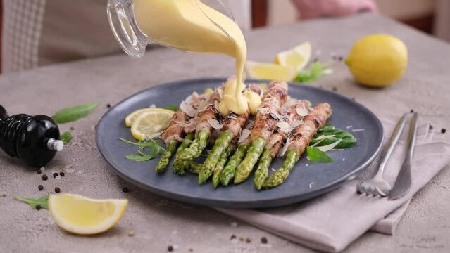 pouring hollandaise sauce onto freshly cooked asparagus wrapped with bacon on ceramic plate