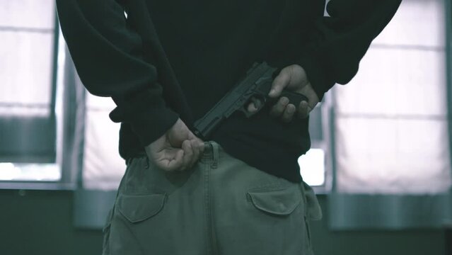 The male assassin put the gun in the rim of his pants, covered his hood, criminal man walking carefully over to look outside his eyes to see his target.