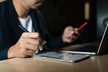 Business planning, E learning concept. Man hand with pen writing on paper notebook while working on laptop computer at home office, close up, dark tone. Student lecture while attending online course