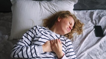 Caucasian woman suffering from heart ache sitting on bed in bedroom after wake up feeling extremely bad. Young adult female experiencing severe pain in her chest. Girl suffers from sudden heart pain