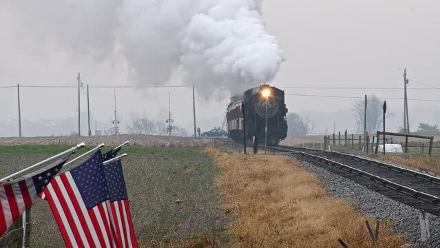 Long View of a Steam Passenger Train Approaching Blowing Smoke and Steam With American Flags on the Fence