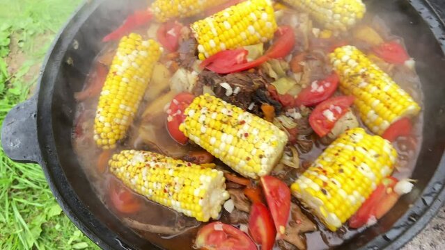 Corn with beef and vegetables. Shurpa in a cauldron. Tomato, red pepper and lamb soup cooked on hot coals. Hot beef hunting goulash or bograch soup with corn cobs, paprika, vegetables and spices.