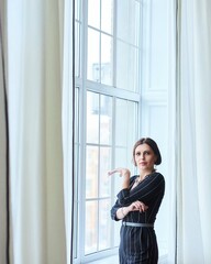A stylish middle-aged woman in dark clothes stands at the window in a bright room. Style and fashion.